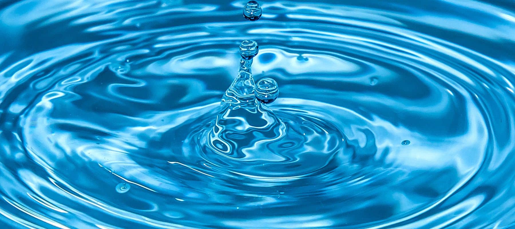 closeup of very blue water with a large water drop falling into it and causing ripples in the water