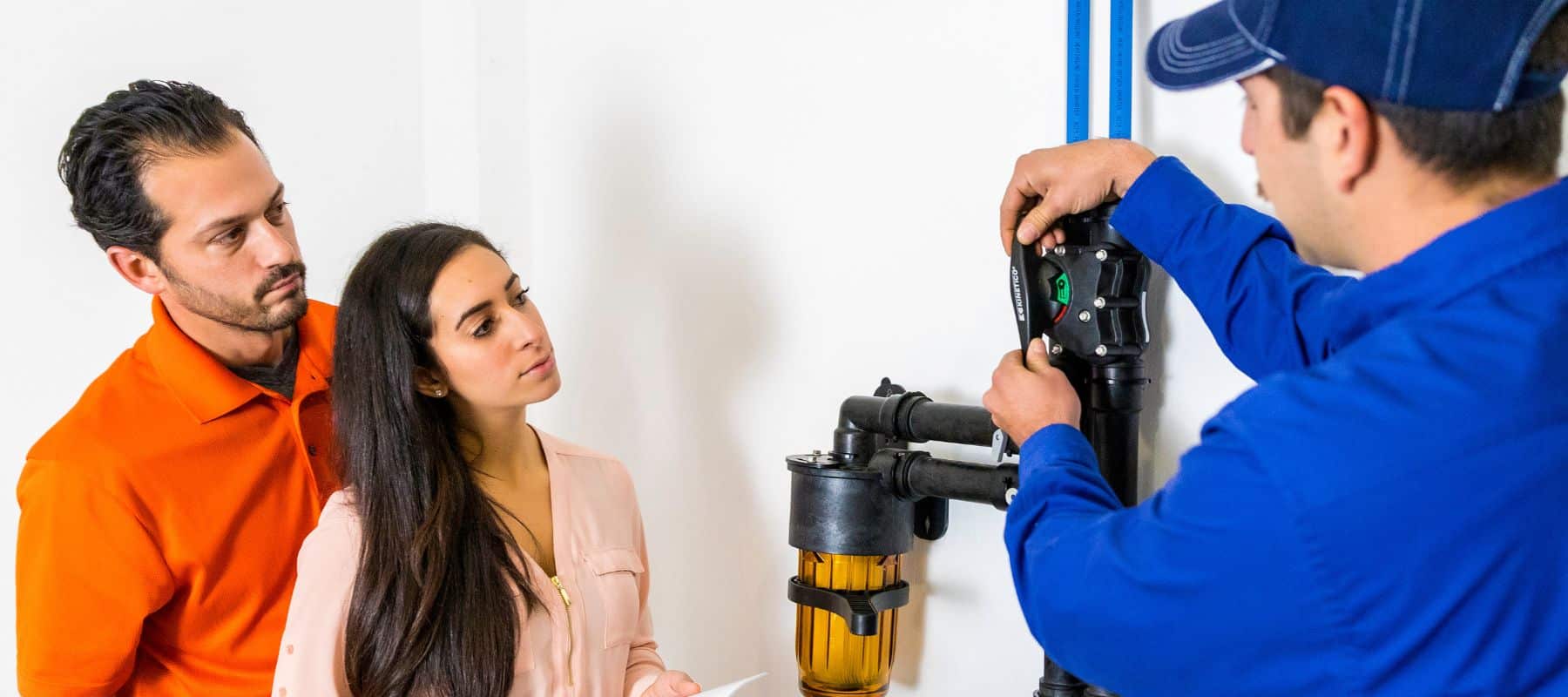 A technician in a blue uniform and cap is explaining a water filtration system to a couple standing nearby. The man, wearing an orange shirt, and the woman, with long dark hair and a light pink blouse, are attentively listening. The technician is adjusting a black component of the filtration system, which includes an orange filter and various pipes, mounted against a white wall